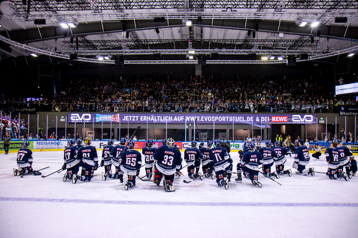 EVO SPORTS FUEL als Partner im Profisport: Eishockeyteam Kasseler Huskies kniet nach dem Spiel auf dem Eis vor voller Tribüne und bedankt sich bei den Fans im Stadion. Auf der Banderole ivosportsfuel.com